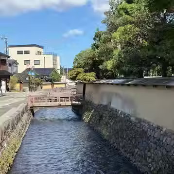A narrow canal runs beside a street and a long pale wall beneath trees in Nagamachi.