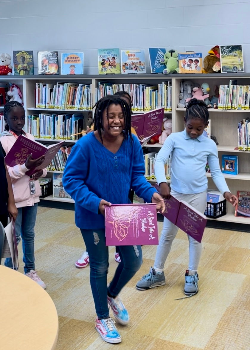Young girl smiling and laughing while holding A Seed Into A FlowHer Workbook in a classroom setting, surrounded by other girls engaging with the workbook