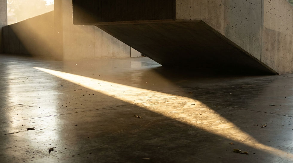 Abstract lighting study. A deep architectural overhang casts a sharp, diagonal line of dark shadow across a bright, sunlit polished concrete floor. Dust motes dance in the light.