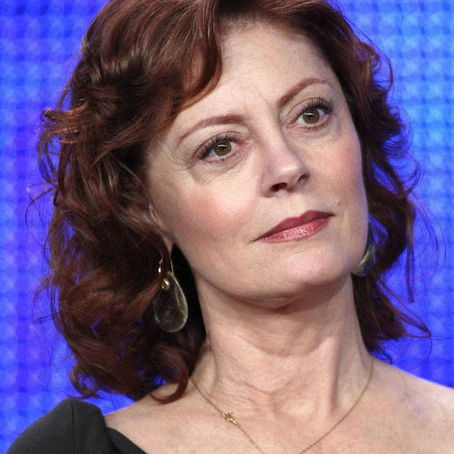 Portrait en couleur de Susan Sarandon, actrice et militante américaine, au regard doux et déterminé, portant une robe noire, des boucles d’oreilles dorées et un collier fin, sur un fond bleu lumineux.