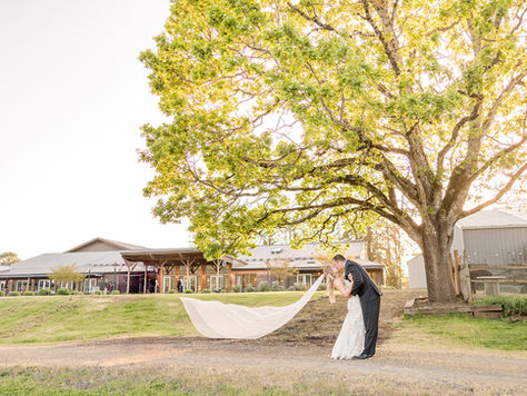 Bride and groom kissing in front of the Abbey Road Farm venue building, with the bride’s veil blowing in the wind on a sunny day.