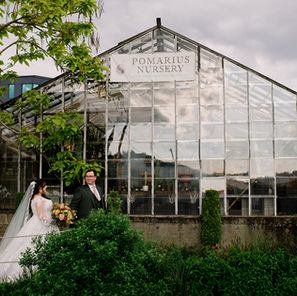 Bride and groom standing in front of the Pomarius Nursery greenhouse at Blockhouse Portland, a lush urban wedding venue in NW Portland. The bride wears a long-sleeved lace gown and carries an organic floral bouquet of terracotta and apricot blooms.
