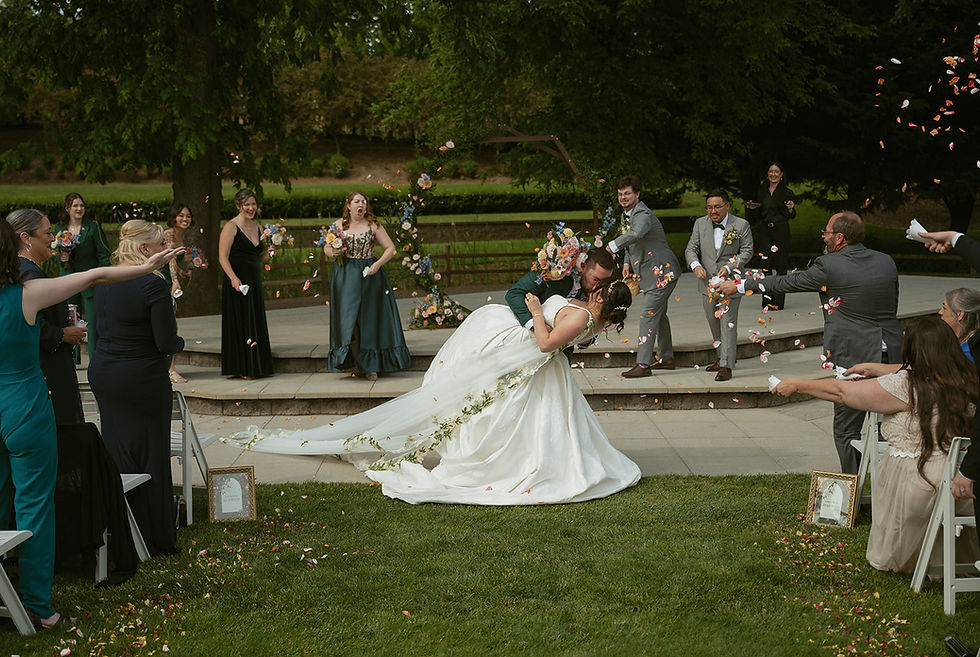Bride and groom share a dramatic dip and kiss during their Aurora Vineyards ceremony as colorful flower petals shower around them, capturing the whimsical, emerald green botanical wedding aesthetic.