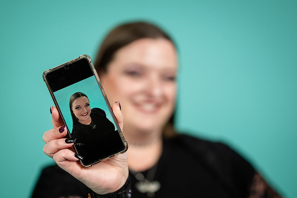 Flick Hamnett-Day The Social Dragonfly is holding a smartphone toward the camera, smiling, with the phone screen showing a clear portrait of herself against a teal background. The woman is out of focus in the background, while the phone and portrait are in sharp focus.