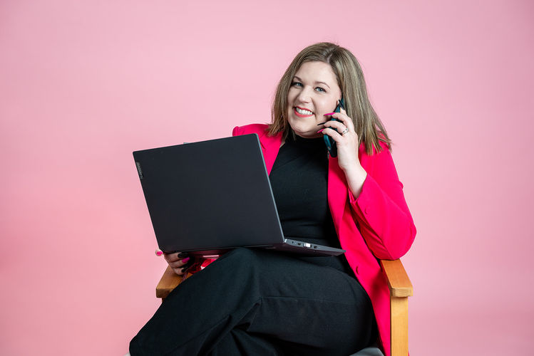 Amie-Leigh from Shh PR smiling during a phone call, holding a laptop and wearing a bold pink blazer against a pink backdrop.