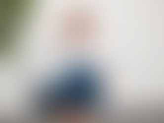 A woman with blonde hair wearing a white shirt and wide-leg blue trousers crouches casually against a white wall, smiling warmly at the camera. A green houseplant is visible on the left side of the frame.