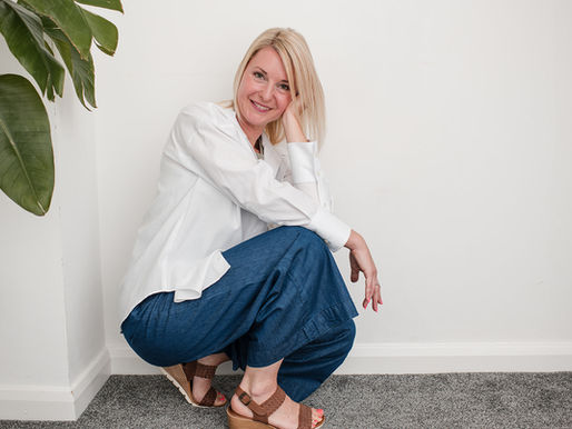 A woman with blonde hair wearing a white shirt and wide-leg blue trousers crouches casually against a white wall, smiling warmly at the camera. A green houseplant is visible on the left side of the frame.