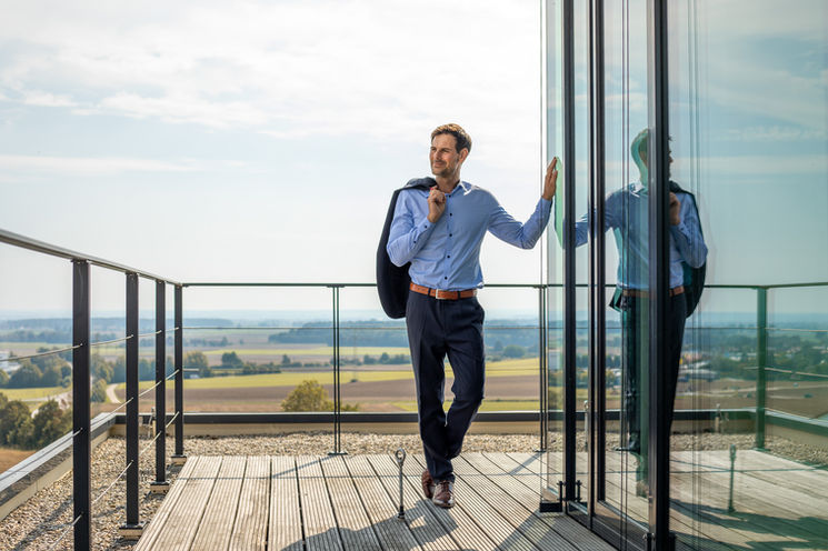 Mann in Hemd auf Balkon blickt lächelnd über weite Landschaft.