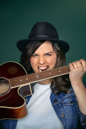 Künstler Portrait: Eine Frau mit schwarzem Hut beißt in den Griff ihrer Gitarre.