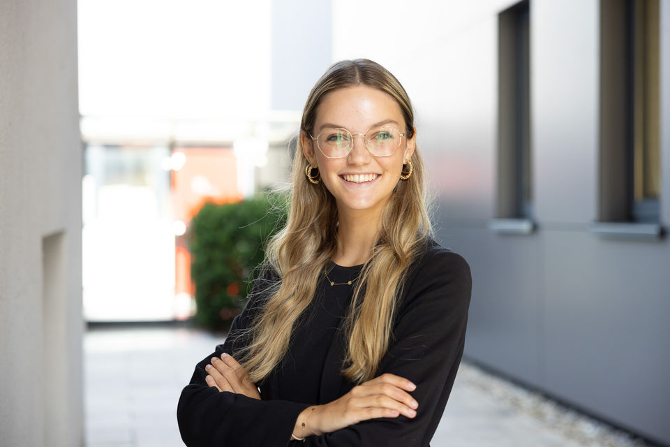 Junge Frau mit Brille lächelt freundlich, Arme verschränkt, im Freien.