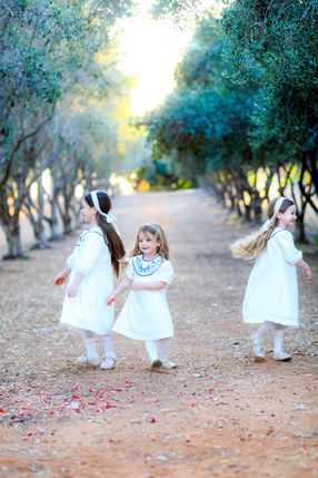 ילדות לבושות לבן הולכות בשביל מוצל בין עצי זית בצילום משפחה בטבע.