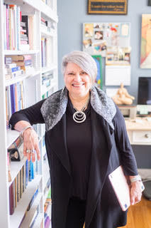 Smiling person with silver hair stands in a cozy office, surrounded by bookshelves. Wearing black attire with statement necklace, holding a tablet.