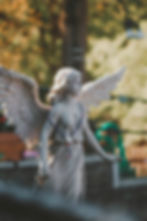 Stone angel statue with detailed wings and rosary stands in a garden, bathed in warm sunlight. Background features blurred green foliage.