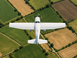 “Top-down aerial view of a white single-engine Cessna 172 flying over patchwork farmland. The airplane is centered in the frame with its wings fully visible and the propeller slightly blurred from motion. Below, geometric fields in varying shades of green and brown are divided by hedgerows, dirt roads, and tree lines under bright daylight with soft shadows.”