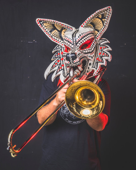 Person wearing a decorative, tribal-style wolf mask plays a trombone against a dark background.