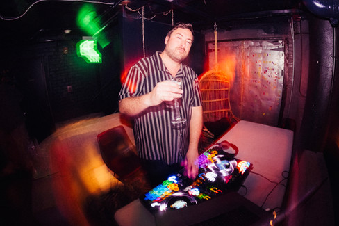 A DJ holding a drink in a dark room; camera light flares