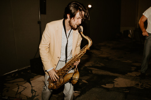 A bearded saxophonist in a light-colored suit and polka dot shirt performs on stage, with another musician partially visible in the background.