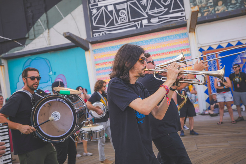 A brass band performs on a wooden boardwalk in front of historic buildings, with musicians playing trumpet, bass drum, and trombone while pedestrians walk by in the background.