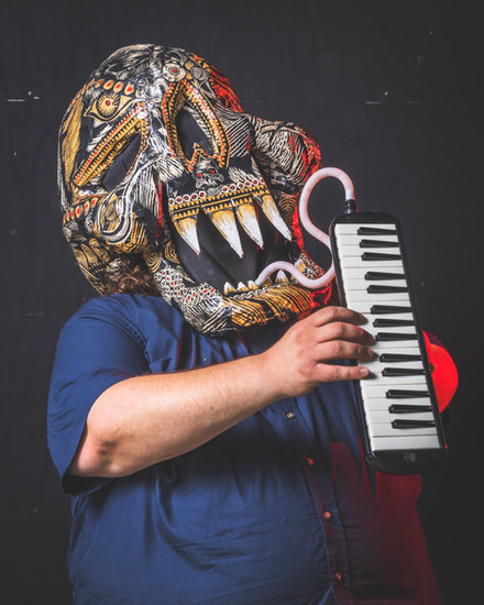 Person wearing a decorative, tribal-style skull mask plays a melodica against a dark background.