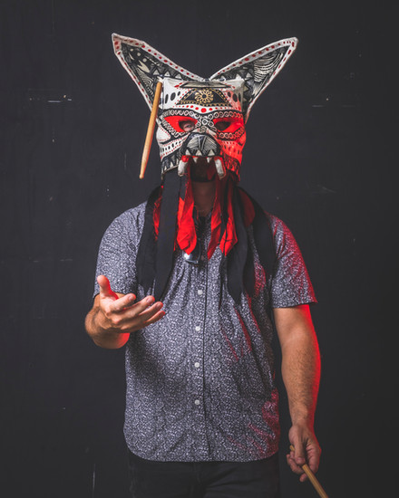 A person in a blue patterned shirt wears an elaborate mask with pointed ears, red and black decorative elements, and dangling tassels, while throwing a drumstick in the air against a dark background.