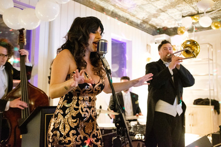 A woman in a sparkly black and gold dress sings into a vintage microphone, with a bassist and trombonist playing in the background. White and gold balloons adorn the ceiling.