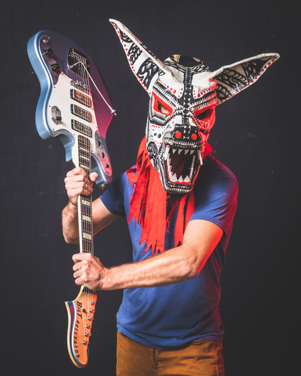Person wearing a decorative, tribal-style horse mask holds a guitar upside-down against a dark background.