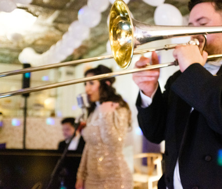 A musician plays a trombone in the foreground, with a woman singing into a microphone and white balloons in the background.