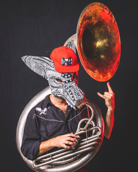 A person wearing a patterned face covering and red "Duff" cap plays a large brass sousaphone, with the instrument's bell glowing in fiery orange and red tones against a dark background.