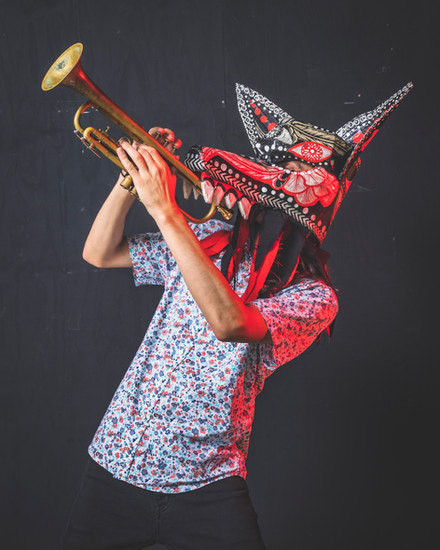 A person in a speckled shirt wears a dramatic horned mask with red, black, and white patterns while playing a brass trumpet against a dark background.