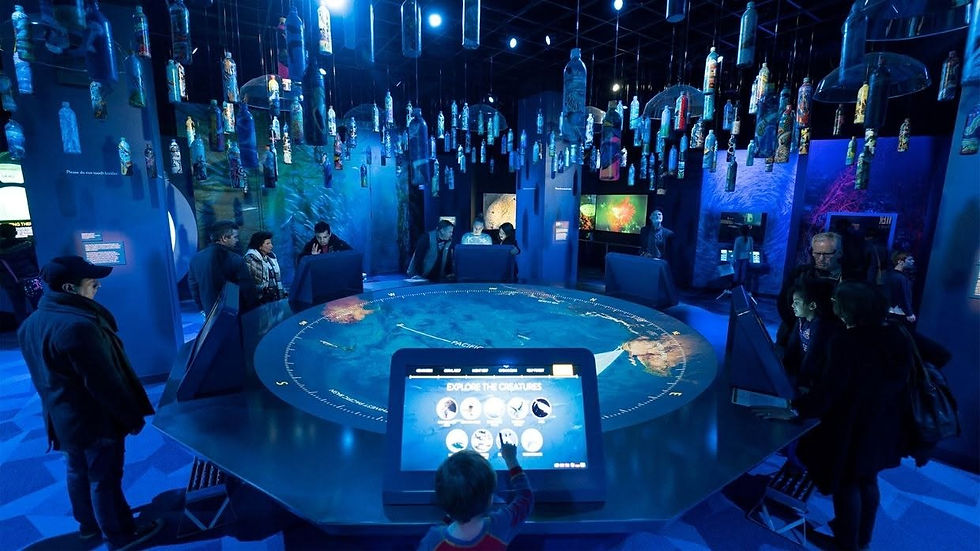 Visitors around a round ocean exhibit in a blue-lit museum, with a screen reading Explore the Creatures and bottles hanging overhead.