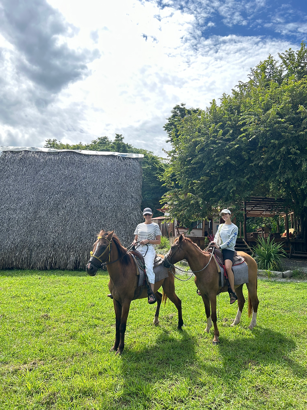 Two smiling riders sit on brown horses in a grassy tropical yard under a cloudy blue sky, beside a thatched hut and trees. front of the tipi of La Chantin in PLaya Guanico