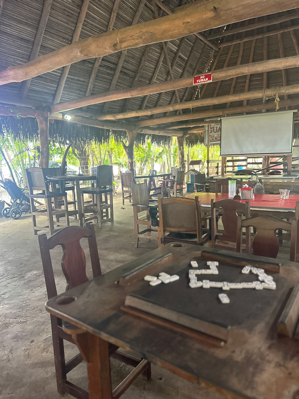 Dominoes on a wooden table in an open-air rustic restaurant with empty chairs, a projector screen, and a red NO FUMAR sign.