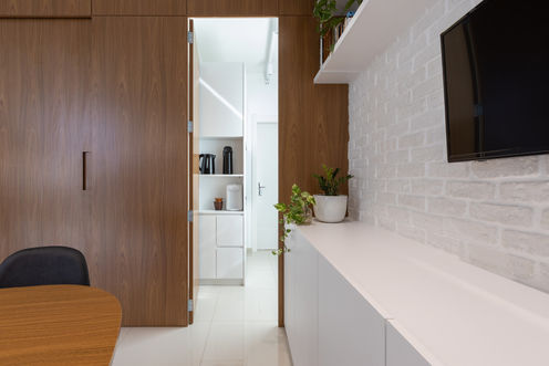 Em primeiro plano, móvel, TV e mesa reuniões. Ao fundo, a copa, móvel com eletrodomésticos sobre prateleiras em nichos, visualizado através de porta aberta embutida em painel de madeira.