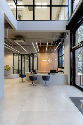 Hall entrada. À direita, porta de aço e vidro. Ao centro, cadeiras giratórias clientes, balcão de atendimento curvo. Ao fundo, painel amadeirado com logomarca, portas de vidro mais à esquerda. Iluminação em linhas. Vista parcial do piso superior.