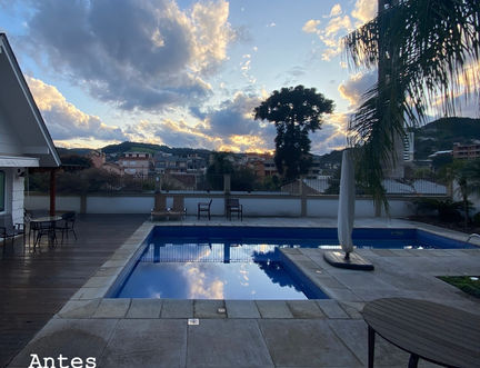 Antes. Piscina em primeiro plano. Salão de festas e deck à esquerda. Ao fundo, muro com grade. Paisagem cidade mais ao fundo. Vegetação à direita.