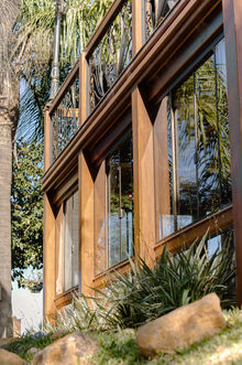 Aproximada estrutura de madeira do anexo frontal. Fechamento em vidro. Varanda superior com guarda-corpo de madeira e detalhes em ferro. Paisagismo em primeiro plano.