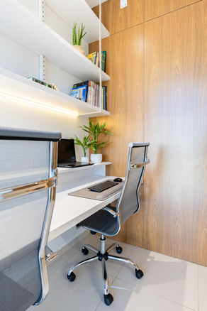 Aproximada. À esquerda, cadeiras de escritório, mesa de trabalho, prateleiras e decoração junto à parede. À direita, painel amadeirado.