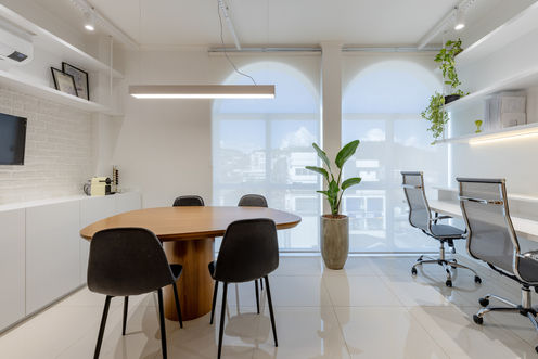 Área de reuniões. Mesa de madeira, cantos arredondados, junto à quatro cadeiras pretas. Ao fundo, parede branca, janela e cortina branca. À esquerda, móvel, prateleiras e parede brancos. À direita, cadeiras de escritório, mesa e prateleiras brancas junto à parede.