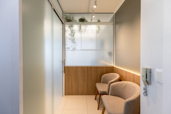 Sala de espera. Imagem aproximada. Cadeiras à direita. Portas de vidro branco fosco à esquerda. Plantas vistas através de vidro fosco ao centro e fundo.