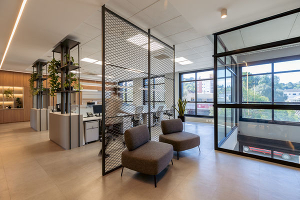 Hall segundo piso. Poltronas junto a uma divisória de ferro. Ao fundo, mesas e computadores. Móvel com prateleiras e plantas entre conjunto de mesas e corredor.