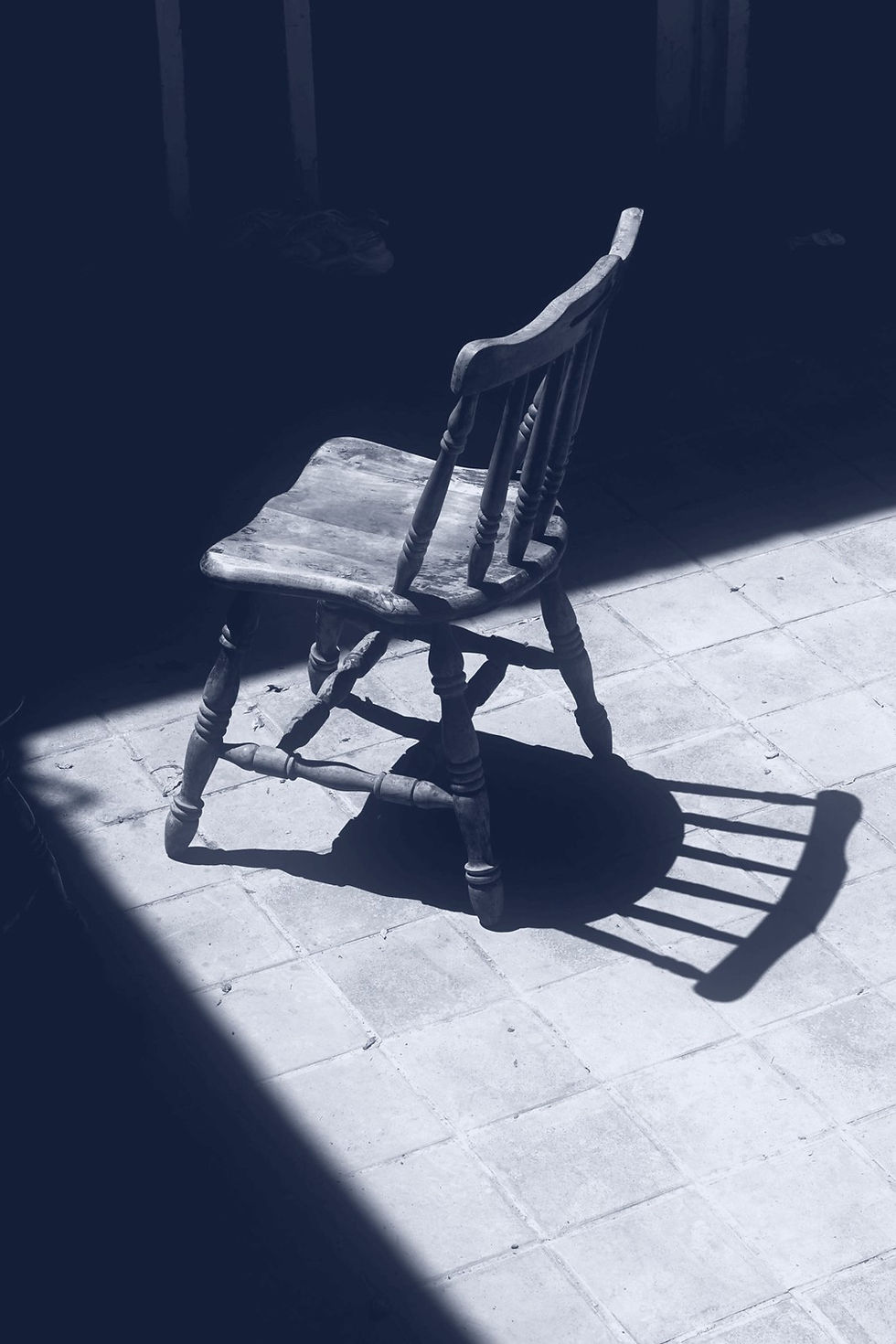 A single worn wooden chair sitting at an angle in a patch of sunlight in a dark room