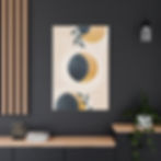 A framed abstract artwork featuring a celestial-inspired design with overlapping circles in shades of deep blue and golden yellow, resembling moon phases. The composition is set against a soft beige background with subtle white specks, evoking a dreamy, cosmic feel. Delicate green leaves extend into the frame, adding an organic touch. The artwork is displayed in a modern interior with a white chair and a blue marbled notebook on the table below, enhancing the minimalist aesthetic