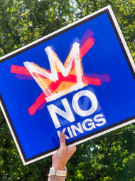 Hand holds a blue sign with a red crossed-out crown and "NO KINGS" text. Green foliage in background, conveying a protest vibe.