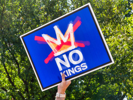 Hand holds a blue sign with a red crossed-out crown and "NO KINGS" text. Green foliage in background, conveying a protest vibe.