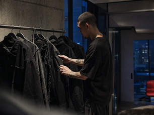 Man comparing black jackets on a garment rack in an indigo-lit Los Angeles apartment