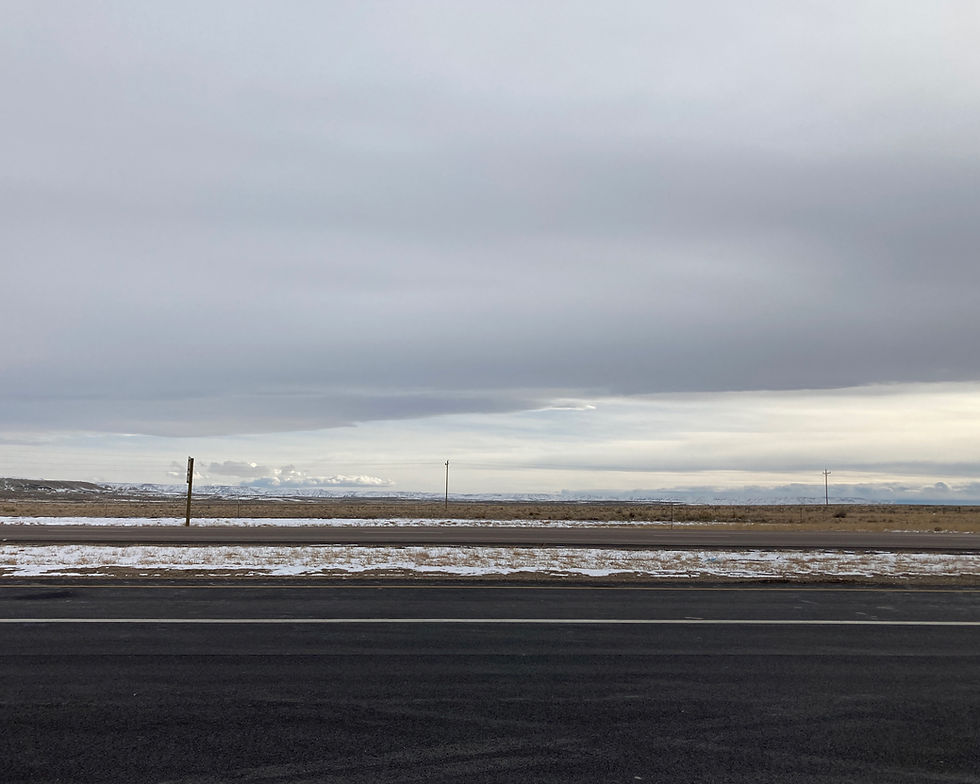 Empty stretch of Interstate 80 / I-80 in Wyoming. The sky is grey, a patchy layer of snow is on the road median, and the Rockie Mountains are in the distant background.