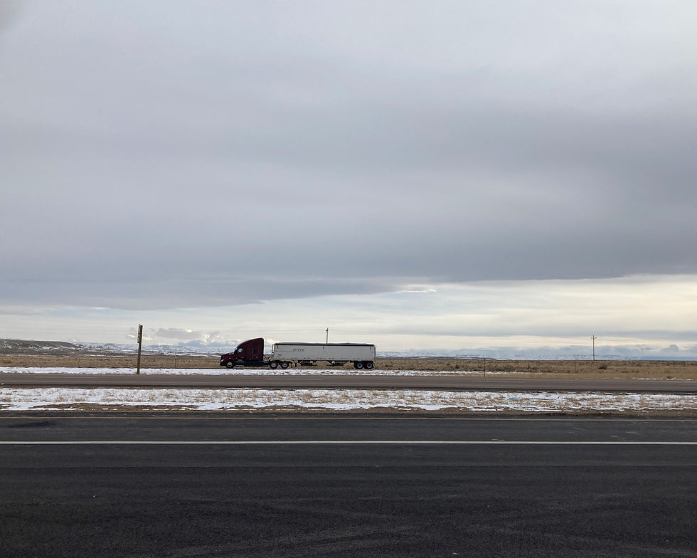 Semi-truck with a red cab and white trailer on an empty stretch of Interstate 80 / I-80 in Wyoming. The sky is grey, a patchy layer of snow is on the road median, and the Rockie Mountains are in the distant background.