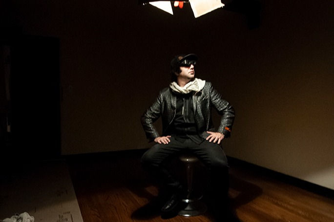 Ari Joshua on a stool under a studio light in a dimly lit room
