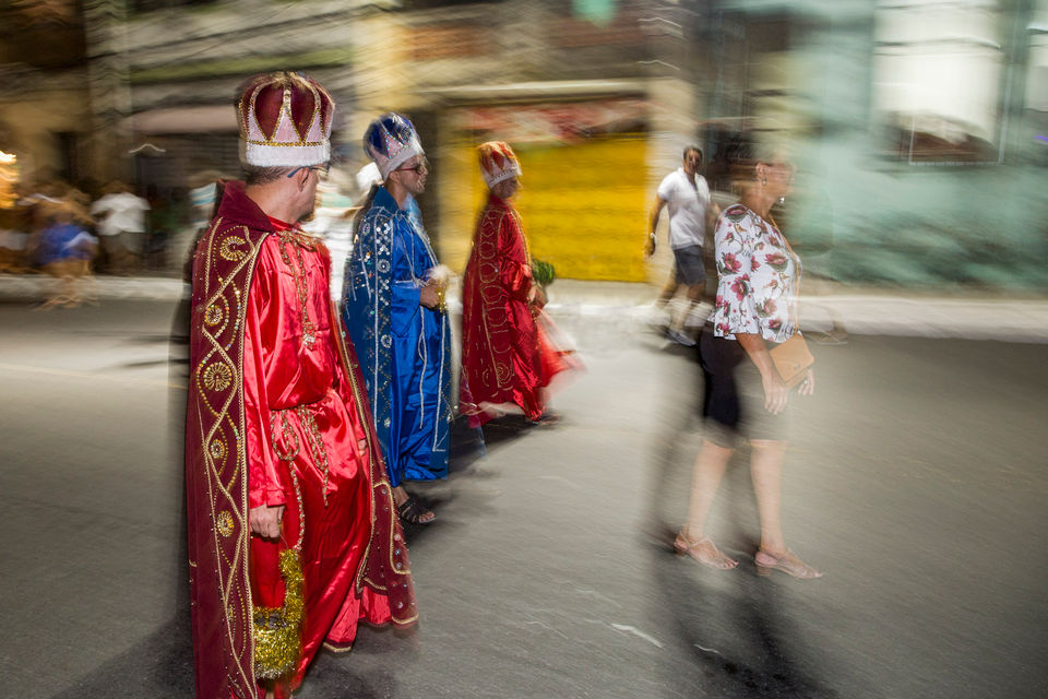 3 homens vestidos de Reis Magos desfilam pela rua. O homem que está mais perto da câmera, assim como o que está mais longe, usam túnicas vermelhas, capas e coroas vermelhas com bordados dourados. O que está ao centro, vem com uma roupa igual, mas toda em tons de azul.