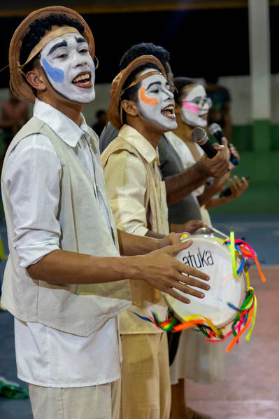 Dois rapazes e uma moça estão alinhados em uma fila diagonal, que vai da esquerda para a direita. Eles vestem roupas claras, com calça, camisa e colete. Têm os rostos pintados de branco e usam chapeu de couro à moda nordestina.
O moço que está em primeiro plano toca um pandeiro enfeitado com fitas coloridas. Os outros dois, cantam ao microfone.